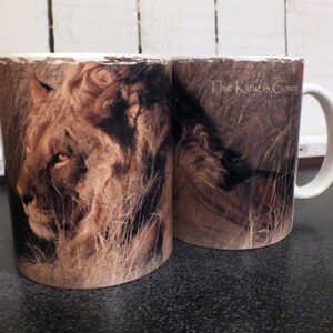 May include: Two ceramic coffee mugs with a lion image. The mug on the left has a close-up of a lion's face. The mug on the right has a lion lying down with the text "The King is Coming".
