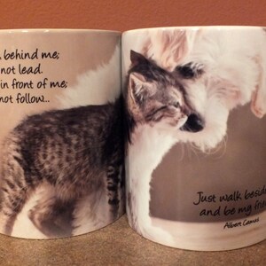 May include: Two white ceramic mugs with handles. One mug features a black and white cat and the text "Don't walk behind me; I may not lead. Don't walk in front of me; I may not follow..." The other mug features a white dog and the text "Just walk beside me and be my friend Albert Camus".