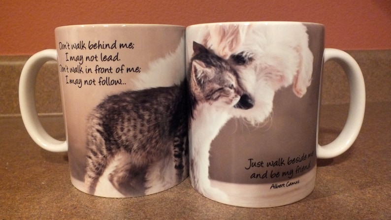 May include: Two white ceramic mugs with handles. One mug features a black and white cat and the text "Don't walk behind me; I may not lead. Don't walk in front of me; I may not follow..." The other mug features a white dog and the text "Just walk beside me and be my friend Albert Camus".