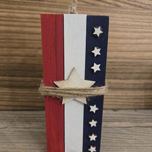 May include: A patriotic wooden decoration in red, white, and blue, resembling a firecracker. The design features painted stripes and star accents, wrapped with twine and a jute loop for hanging. A wooden star is attached to the front.