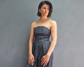 May include: A woman wearing a strapless denim jumpsuit with wide legs and cuffed hems. She is standing in front of a gray wall and has her hair pulled back.