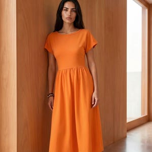 May include: A solid orange midi dress featuring a round neckline, short sleeves, and a fitted bodice that transitions into a gathered, flowing skirt that falls below the knees. The garment is constructed from a smooth, opaque fabric.