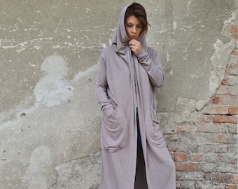 Könnte beinhalten: Ein langer, Kapuzenpullover in hellbrauner Farbe. Der Pullover hat Taschen an den Seiten und eine Kapuze mit Kordelzug. Das Model trägt den Pullover mit einer blauen Jeans und weißen Turnschuhen.