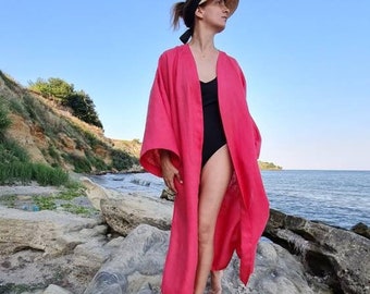 May include: A woman wearing a pink linen robe stands on a large rock formation by the ocean. The robe has long sleeves and a wide, open neckline. The woman is barefoot and has her hair pulled back.