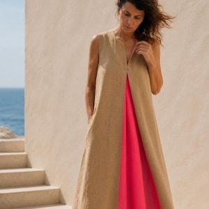 Può includere: Un abito lungo senza maniche con scollo a V. Lo strato esterno è in tessuto marrone chiaro, simile al lino, che rivela un pannello rosa acceso al centro. L'abito ha una vestibilità ampia e tasche.