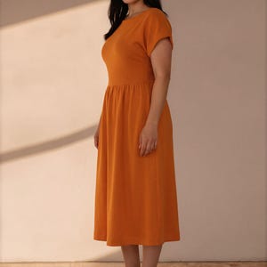 May include: A solid orange midi dress featuring a round neckline, short sleeves, and a gathered waistline that flows into a full skirt. The garment is paired with neutral-toned athletic sneakers, completing a simple and functional ensemble.