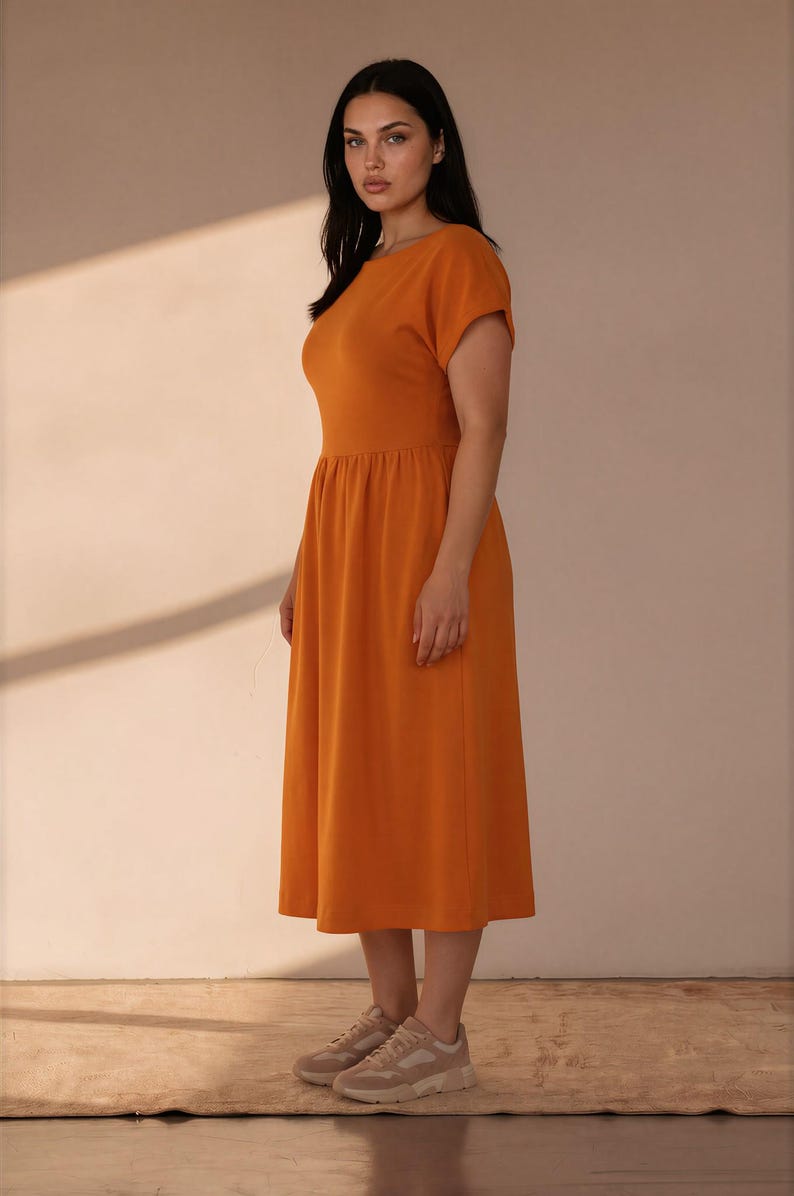 May include: A solid orange midi dress featuring a round neckline, short sleeves, and a gathered waistline that flows into a full skirt. The garment is paired with neutral-toned athletic sneakers, completing a simple and functional ensemble.
