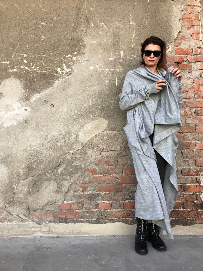 May include: A gray long-sleeved cardigan with a draped design. The cardigan is worn by a person with dark hair and sunglasses. The person is standing in front of a brick wall.