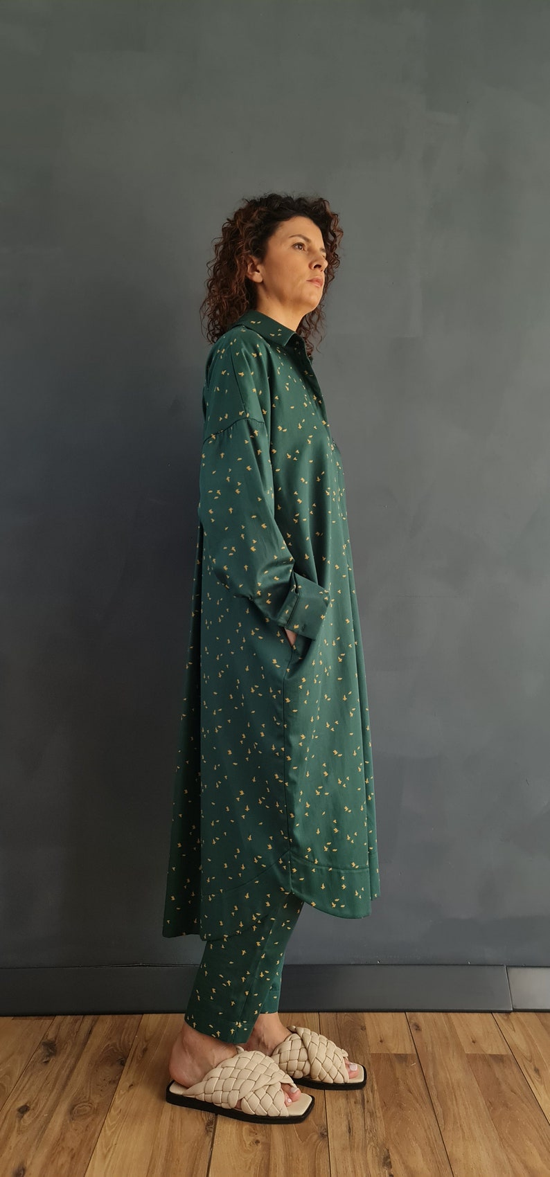 May include: A long, green shirt dress with a small yellow floral pattern. The dress has a collared neckline and long sleeves. The model is wearing the dress with matching pants and beige woven sandals.