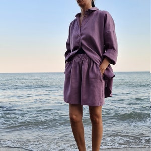 Może przedstawiać: Kobieta w fioletowym komplecie z lnianej koszuli i spodenek stoi na piaszczystej plaży z oceanem w tle. Ma na sobie słomkowy daszek i trzyma ręce w kieszeniach.