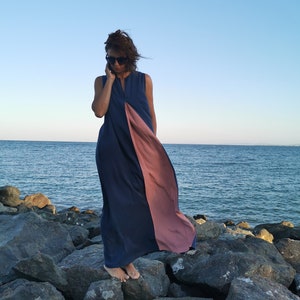 May include: A woman wearing a long, flowing dress with a blue and pink color scheme. The dress has a split in the front, revealing the pink fabric underneath. She is standing on a rocky beach with the ocean in the background.