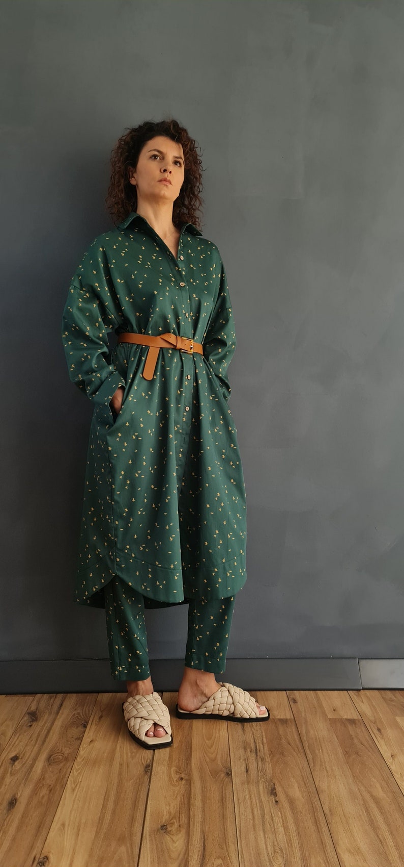 May include: A green shirt dress with a small yellow floral pattern and a brown belt. The dress is worn with matching green pants and beige woven sandals.