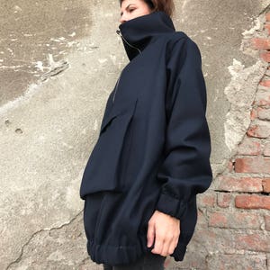 May include: A navy blue oversized jacket with a high collar and a zipper closure. The jacket has a large pocket on the front and a drawstring at the bottom.