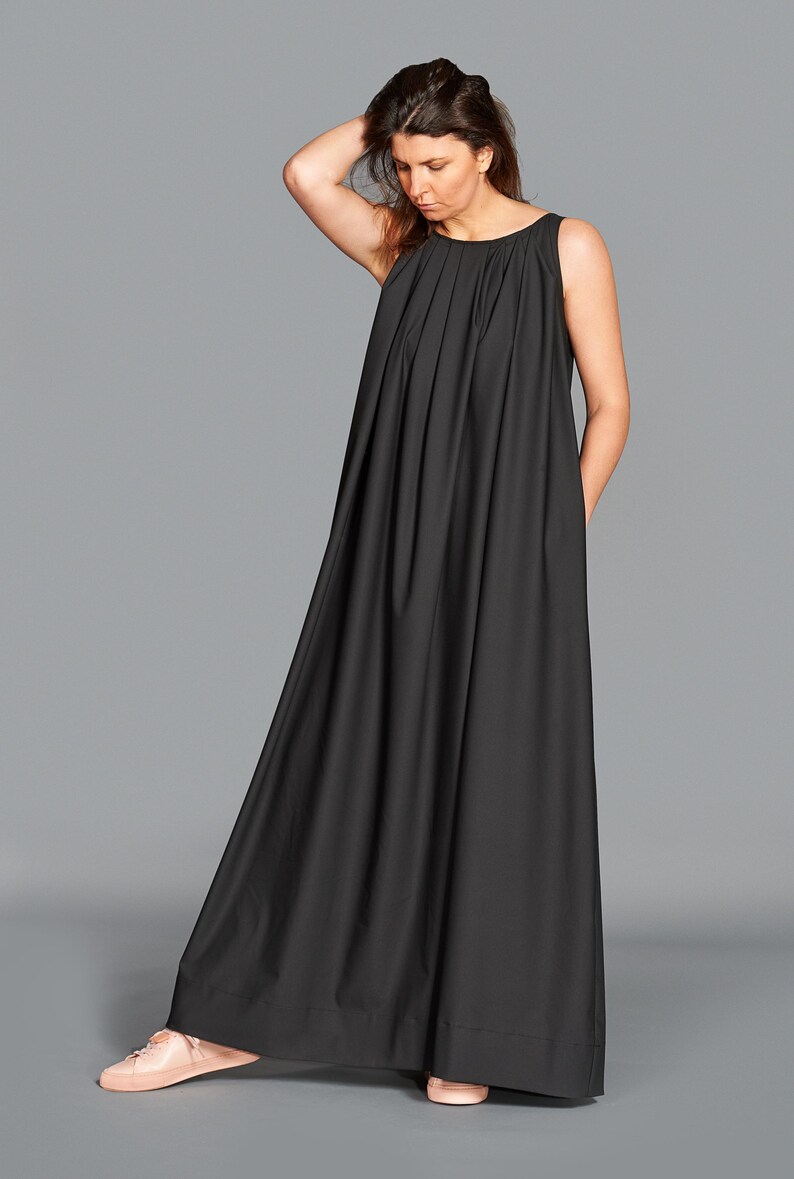 May include: A sleeveless, floor-length black dress with a pleated bodice. The dress has a flowing, A-line skirt and is worn with light pink sneakers. The model is standing against a gray background.
