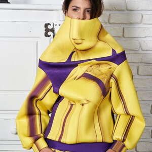 Puede incluir: Una sudadera de rayas amarillas y moradas con un estampado de cara de mujer. La sudadera tiene cuello alto y mangas largas. La mujer lleva pantalones cortos a juego de rayas amarillas y moradas.