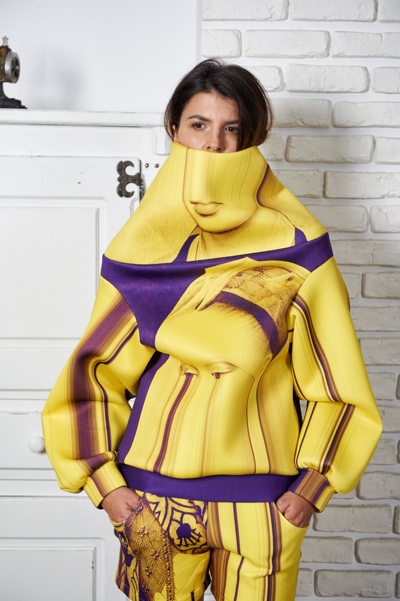 Puede incluir: Una sudadera de rayas amarillas y moradas con un estampado de cara de mujer. La sudadera tiene cuello alto y mangas largas. La mujer lleva pantalones cortos a juego de rayas amarillas y moradas.