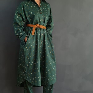 May include: A woman wearing a green and yellow patterned shirt dress with a brown belt and matching pants. She is wearing beige woven sandals.