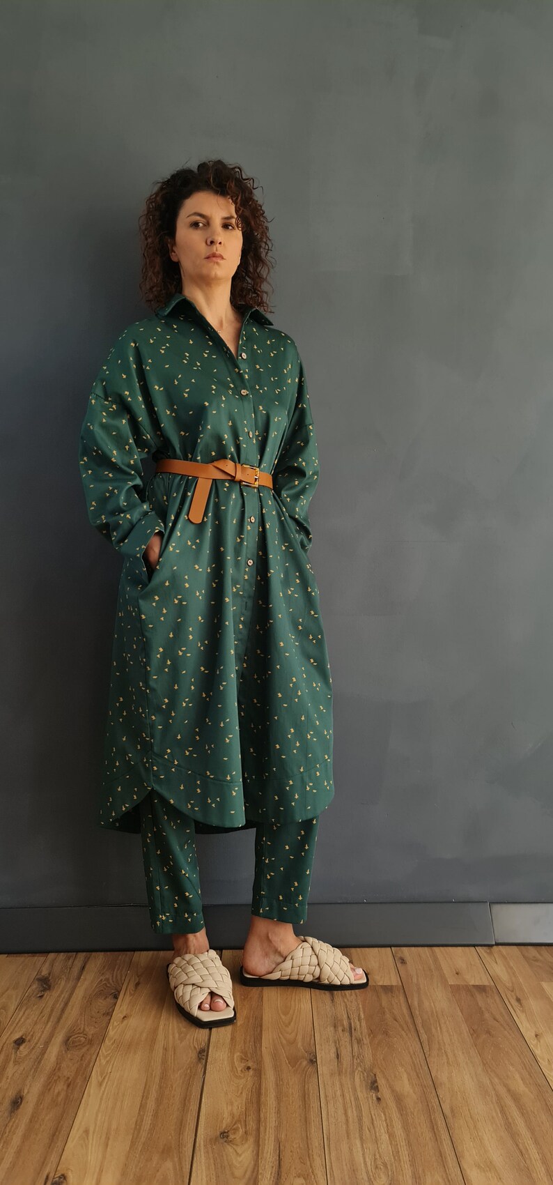 May include: A woman wearing a green and yellow patterned shirt dress with a brown belt and matching pants. She is wearing beige woven sandals.