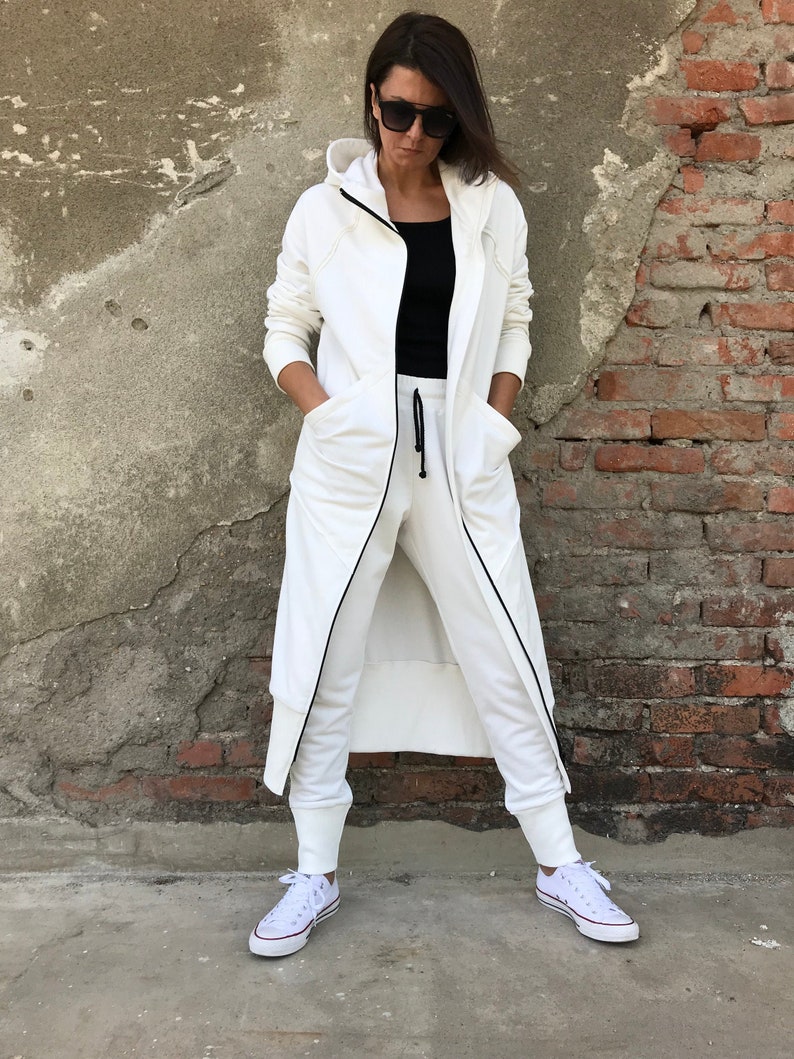 May include: A white hooded long-sleeve jacket with a zipper closure and black trim. The jacket has two pockets and is worn over a black top and white pants.