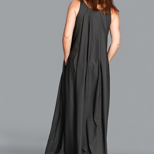May include: A sleeveless, floor-length black dress with a pleated back detail. The dress has a relaxed fit and pockets. The model is wearing the dress, with hands in the pockets, against a gray backdrop.
