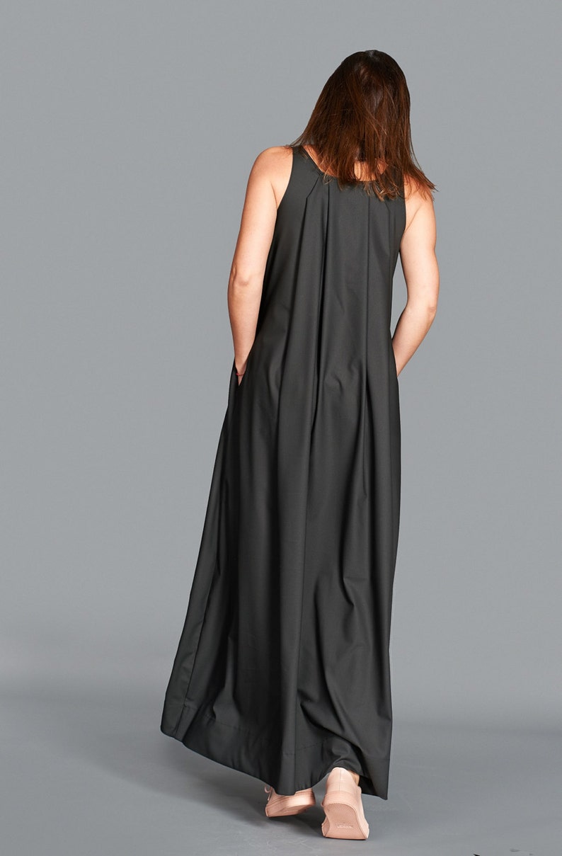 May include: A sleeveless, floor-length black dress with a pleated back detail. The dress has a relaxed fit and pockets. The model is wearing the dress, with hands in the pockets, against a gray backdrop.