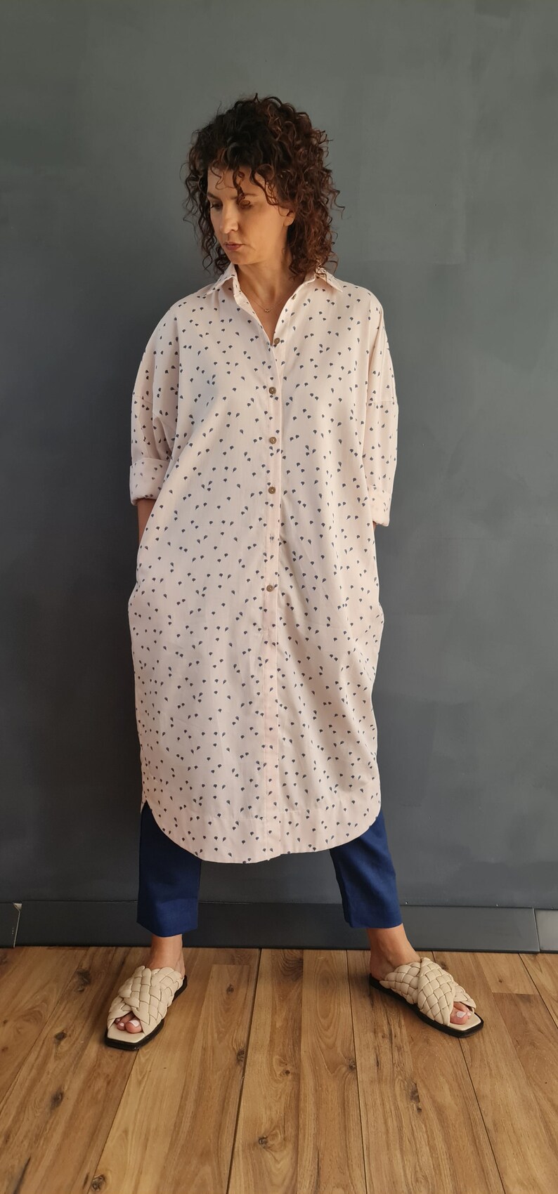 May include: A long-sleeved, pink button-down shirt with a small, dark blue print. The shirt is worn with dark blue pants and beige sandals.