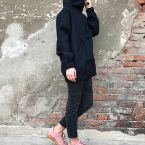 May include: A navy blue oversized jacket with a high collar and a zipper closure. The jacket has a large pocket on the front and a drawstring at the waist. The model is wearing black jeans and pink sneakers.