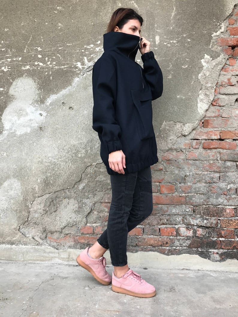 May include: A navy blue oversized jacket with a high collar and a zipper closure. The jacket has a large pocket on the front and a drawstring at the waist. The model is wearing black jeans and pink sneakers.