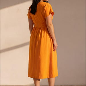 May include: A vibrant orange midi dress featuring short sleeves, a round neckline, and a cinched waistline with a gathered skirt. The dress has a simple, solid-colored design and is paired with beige and white athletic sneakers with thick soles.