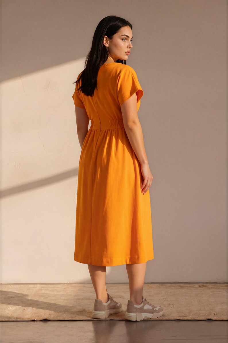 May include: A vibrant orange midi dress featuring short sleeves, a round neckline, and a cinched waistline with a gathered skirt. The dress has a simple, solid-colored design and is paired with beige and white athletic sneakers with thick soles.