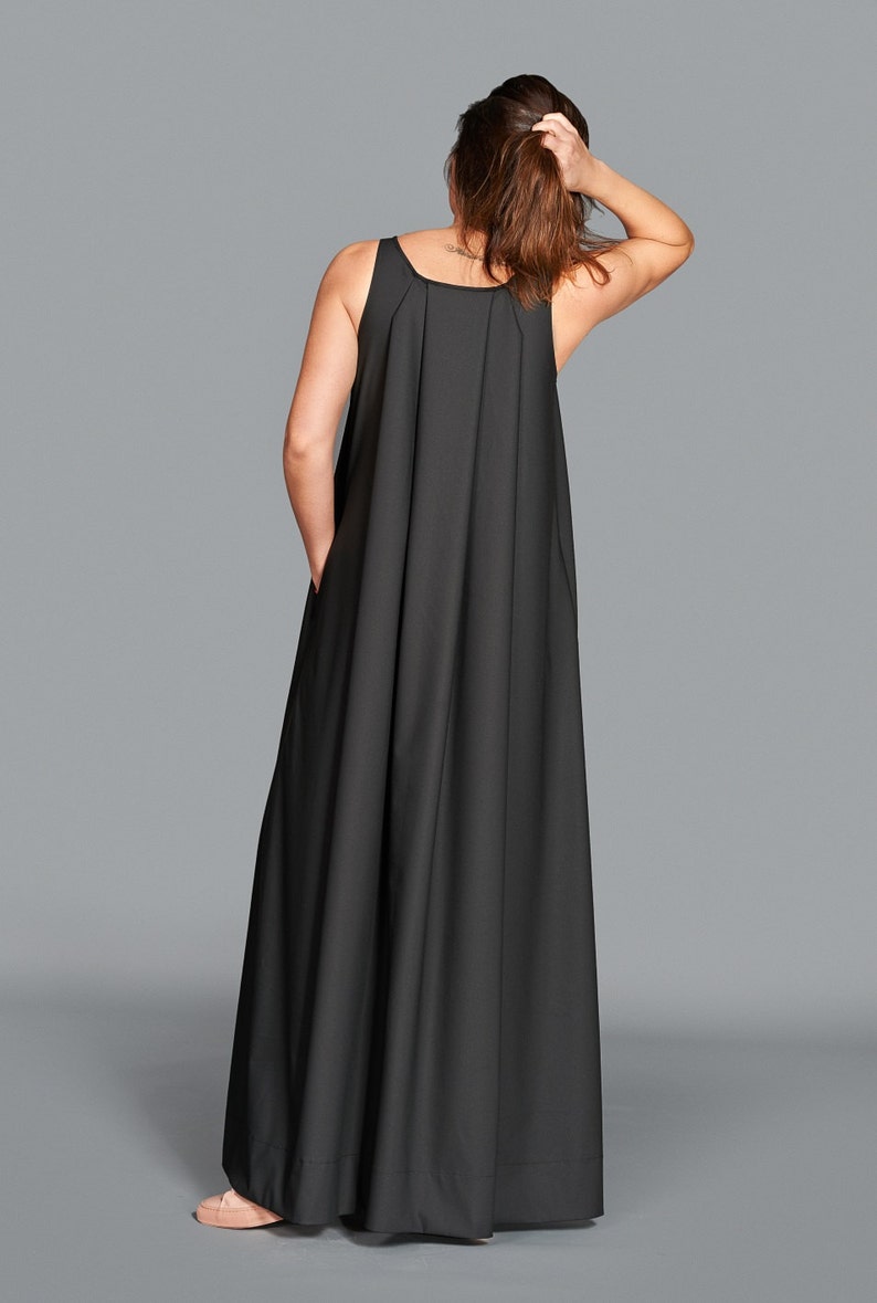 May include: A black sleeveless maxi dress with a pleated bodice and a flowing skirt. The dress has a scoop neckline and pockets. The model is wearing the dress and has their hand in their hair.