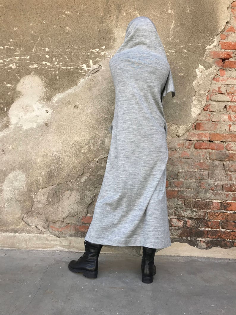 May include: A long, gray, hooded dress with a loose fit. The dress is made of a soft, comfortable fabric and has a relaxed, casual style. The model is wearing black boots.