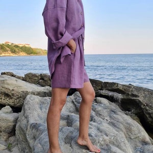Może przedstawiać: Kobieta w komplecie z koszuli i spodenek z lawendowego lnu. Stoi na skalistym brzegu z oceanem w tle.