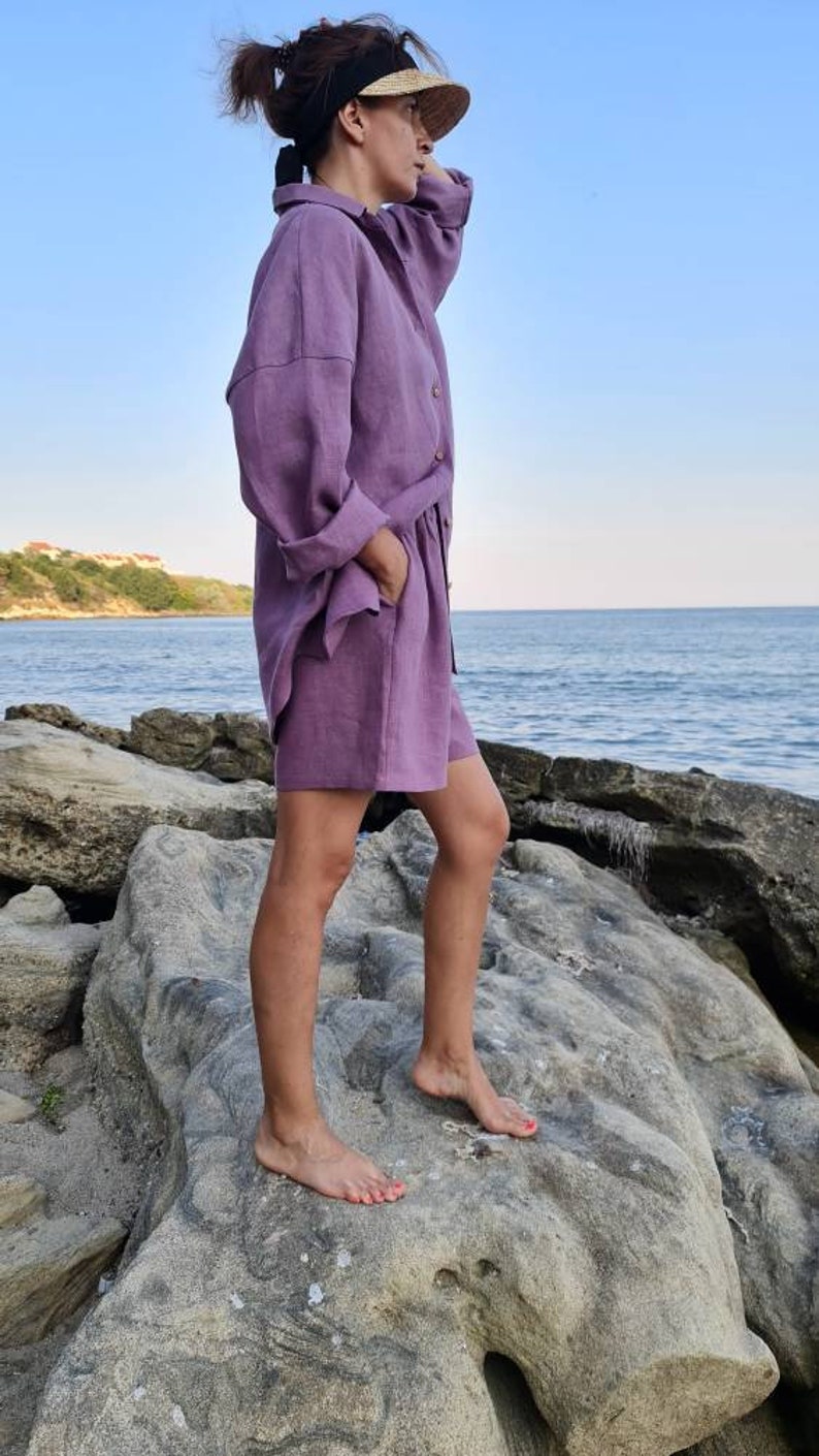 Może przedstawiać: Kobieta w komplecie z koszuli i spodenek z lawendowego lnu. Stoi na skalistym brzegu z oceanem w tle.