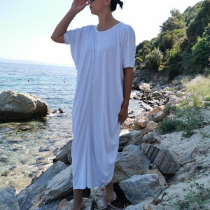 May include: A woman wearing a white, loose-fitting maxi dress with short sleeves stands on rocks by the ocean. She is wearing sunglasses and has her hair pulled back.