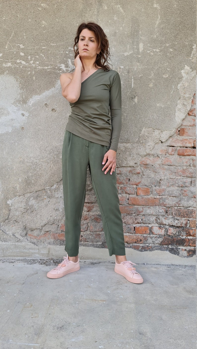 Puede incluir: Una mujer con un top verde oliva de un solo hombro y pantalones verde oliva. Lleva zapatillas rosas.