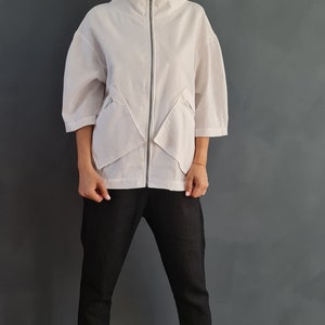 May include: A white zip-up jacket with a high collar and angled pockets. The jacket is worn over black pants and white sneakers.
