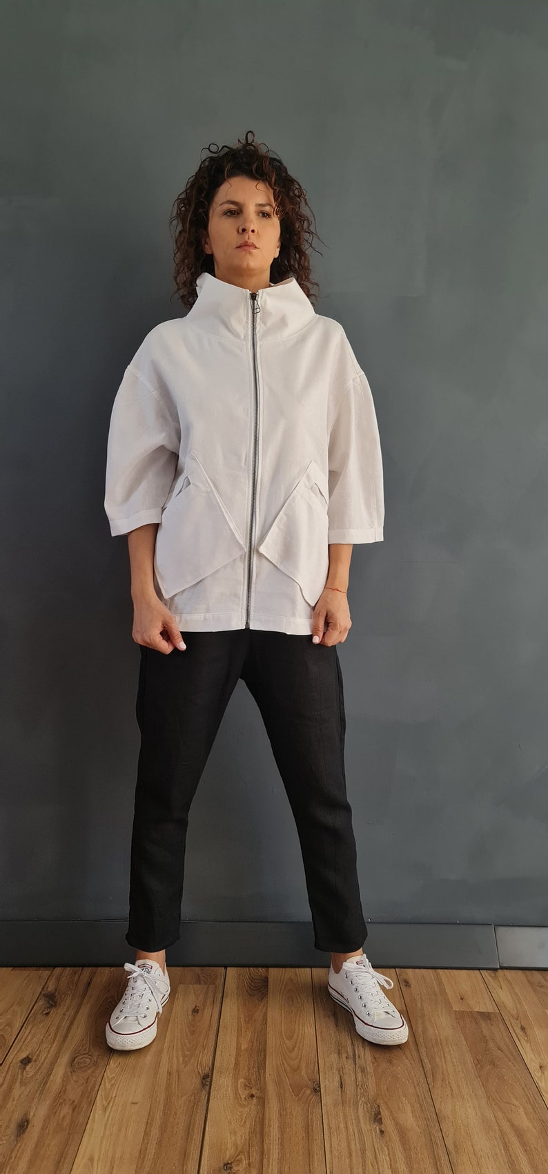 May include: A white zip-up jacket with a high collar and angled pockets. The jacket is worn over black pants and white sneakers.