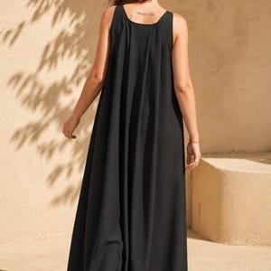 May include: A black sleeveless maxi dress with a flowing, wide-leg design. The dress has a scoop neckline and a loose, comfortable fit. The model is wearing black sandals. The background is a neutral beige color with shadows.