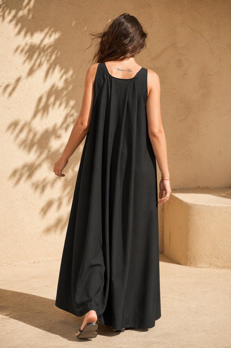 May include: A black sleeveless maxi dress with a flowing, wide-leg design. The dress has a scoop neckline and a loose, comfortable fit. The model is wearing black sandals. The background is a neutral beige color with shadows.