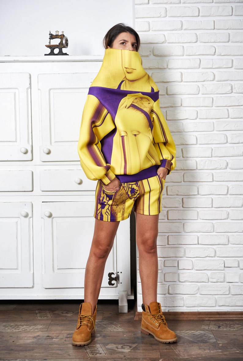 Puede incluir: Una mujer con un jersey amarillo y morado con estampado, cuello alto y pantalones cortos a juego. Lleva botas marrones.