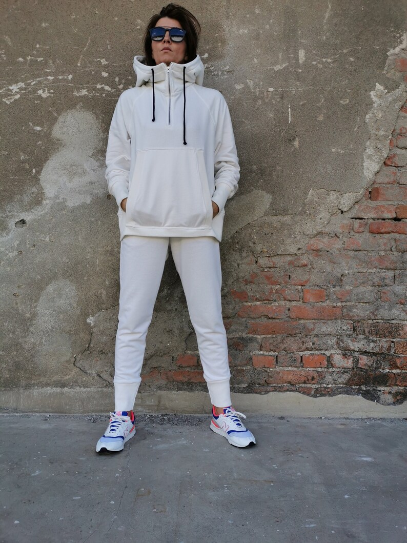 May include: A white hooded sweatshirt with a zipper and a white pair of sweatpants. The person is wearing white sneakers with blue and pink accents.