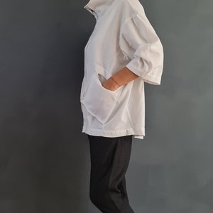 May include: A white collared shirt with a zipper closure and a large pocket on the front. The shirt is worn with black pants and white sneakers.