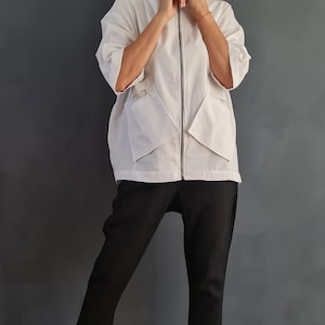 May include: A white zip-up jacket with a high collar and pockets. The jacket is worn over black pants and white sneakers.