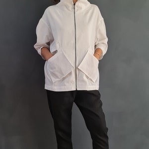 May include: A white zip-up jacket with large pockets and a high collar. The jacket is worn over black pants and white sneakers.