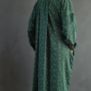 May include: A long, green shirt dress with a small yellow floral pattern. The dress has a collared neckline and long sleeves. The model is wearing the dress with a pair of matching green pants and beige woven sandals.
