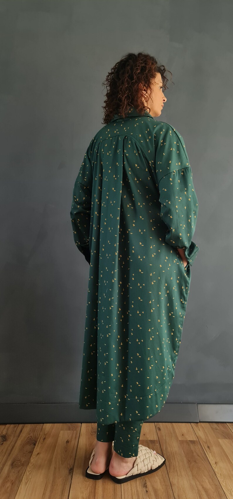 May include: A long, green shirt dress with a small yellow floral pattern. The dress has a collared neckline and long sleeves. The model is wearing the dress with a pair of matching green pants and beige woven sandals.