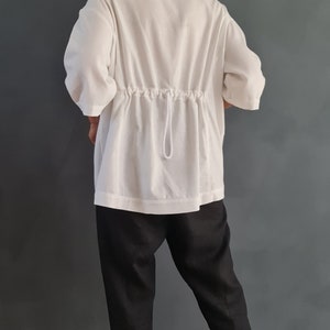 May include: A white, loose-fitting shirt with a drawstring at the waist. The shirt is worn with black pants and white sneakers.