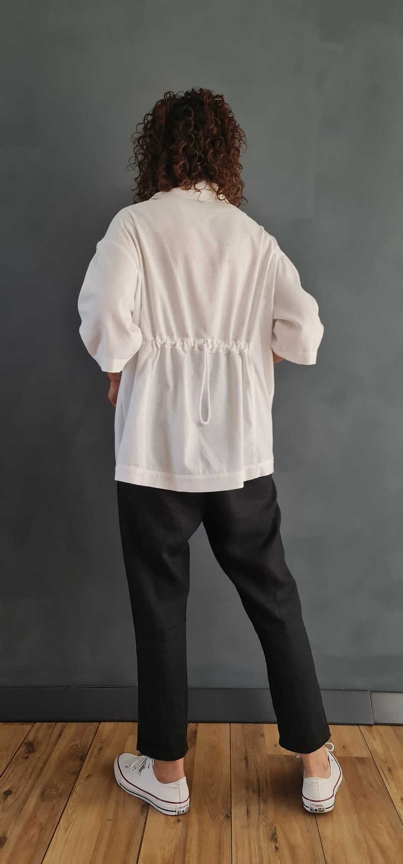 May include: A white, loose-fitting shirt with a drawstring at the waist. The shirt is worn with black pants and white sneakers.