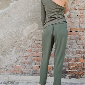 Puede incluir: Una mujer con un top verde oliva de un solo hombro y pantalones verde oliva. Lleva zapatillas rosas y est&aacute; de pie frente a una pared de ladrillo.